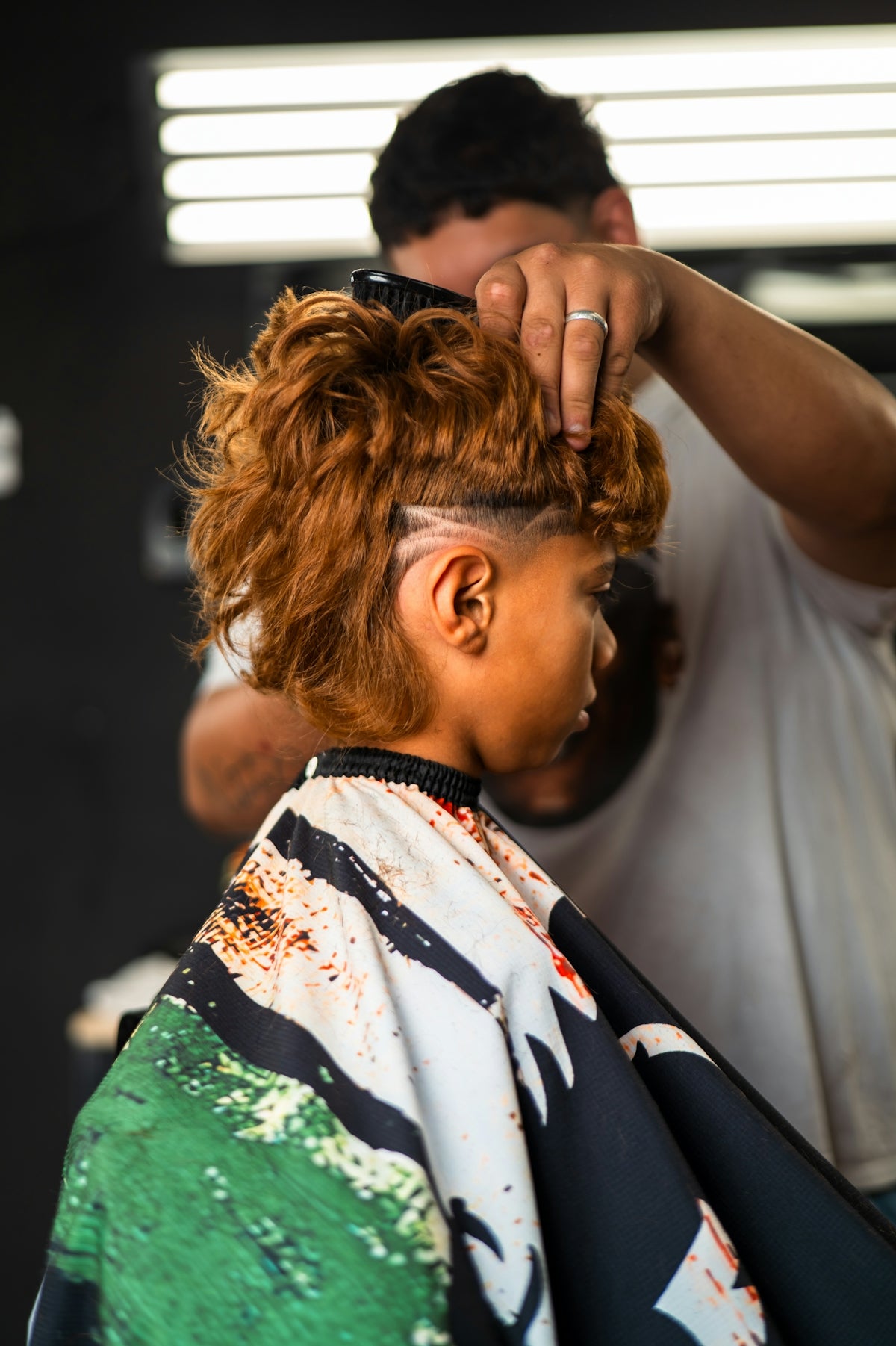 A barber styling a person's curly hair with a shaved design in a salon