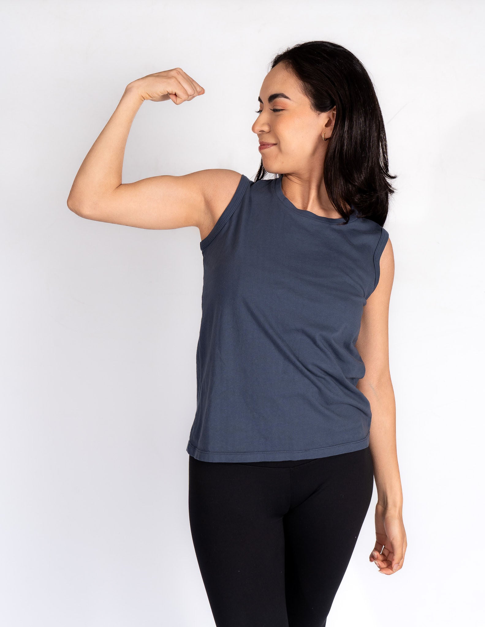 A female model wearing a brown organic cotton muscle tank top with sleeveless design, paired with blue ripped jeans and a wide-brimmed hat.