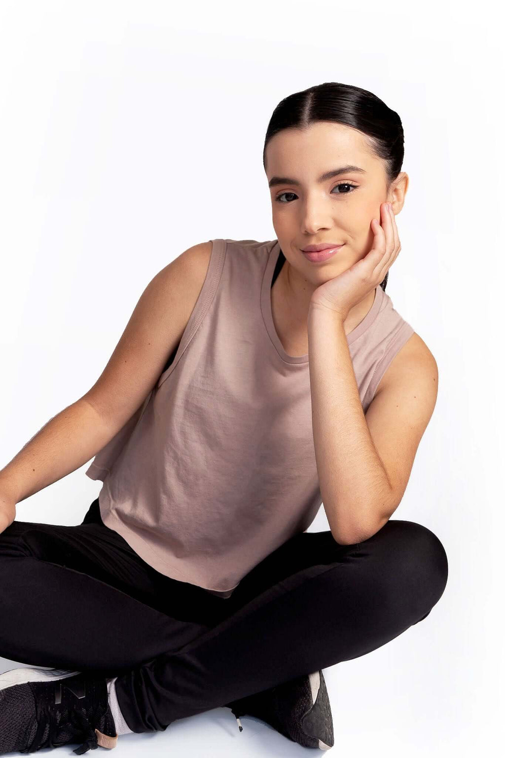 Woman sitting cross-legged on a white background wearing a sleeveless top and black pants.