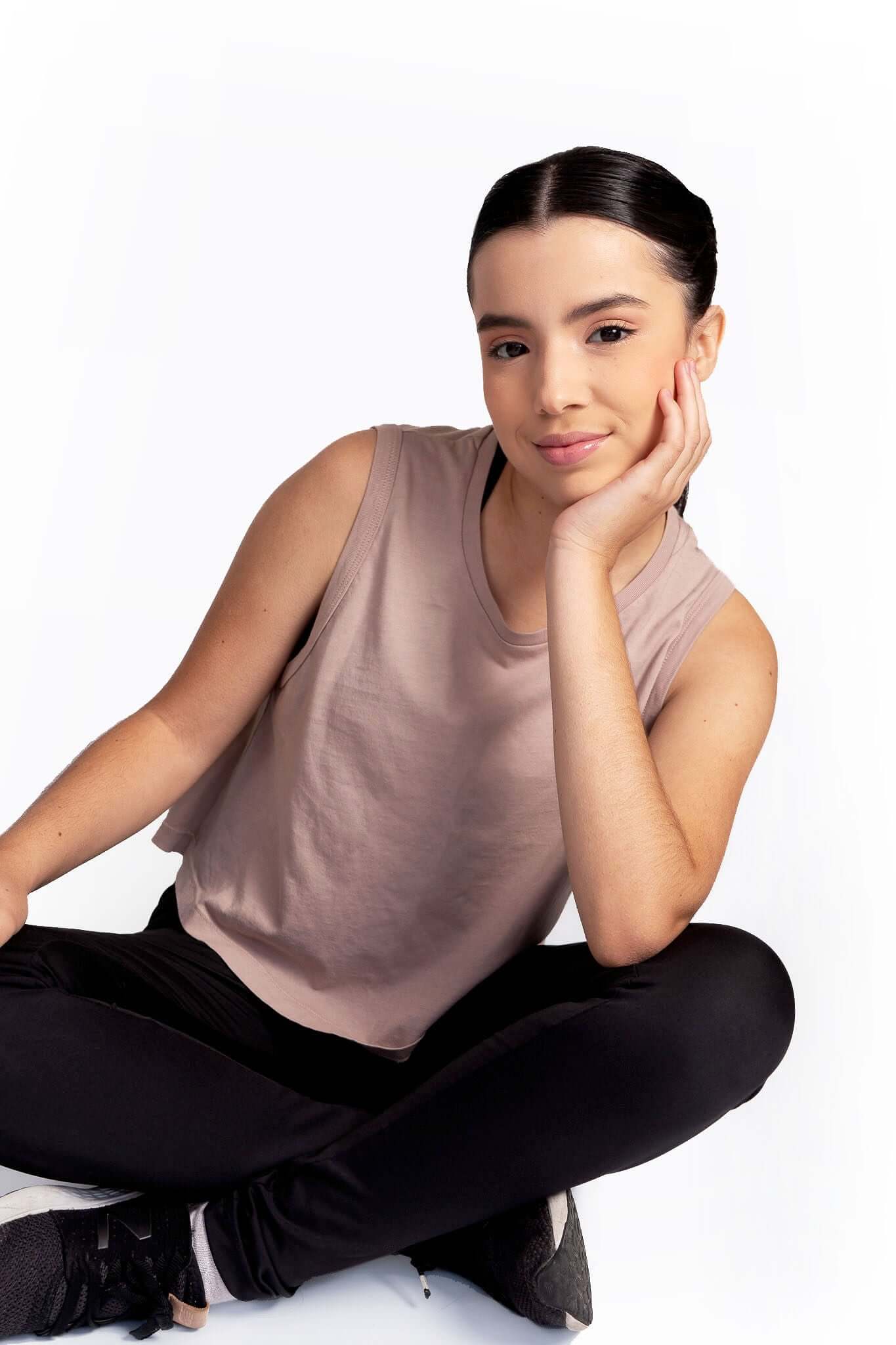 Woman sitting cross-legged on a white background wearing a sleeveless top and black pants.